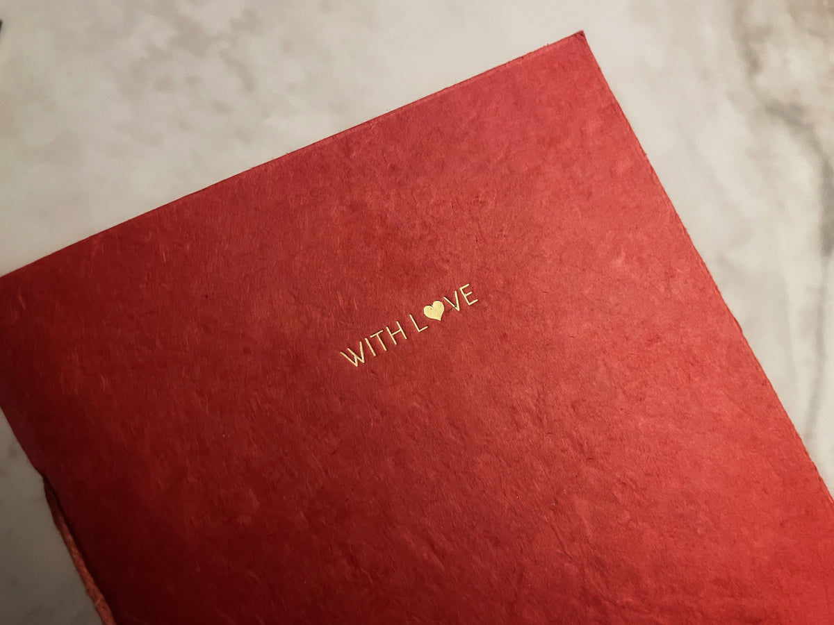 A close-up of a textured red notebook or journal cover with the words 'WITH LOVE' printed in gold foil, resting on a light marble surface.