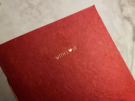 A close-up of a textured red notebook or journal cover with the words 'WITH LOVE' printed in gold foil, resting on a light marble surface.